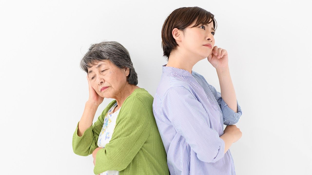 An elderly mother and her adult daughter standing back-to-back, both looking worried.