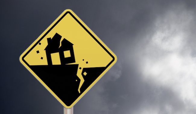 A yellow and black road sign showing a house falling off a cliff, indicating earthquake or landslide risk.