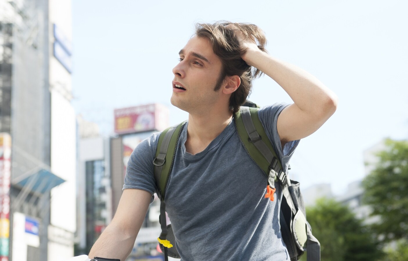 A confused-looking foreign man with a backpack scratching his head on a street in Japan.