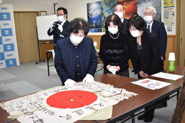 A family wearing masks and white gloves respectfully views a returned Yosegaki Hinomaru flag on a table.