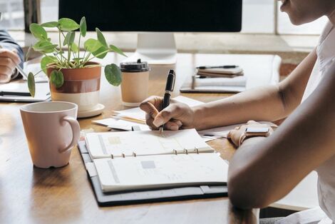 A person writing notes in a notebook at a desk with a computer and plants.