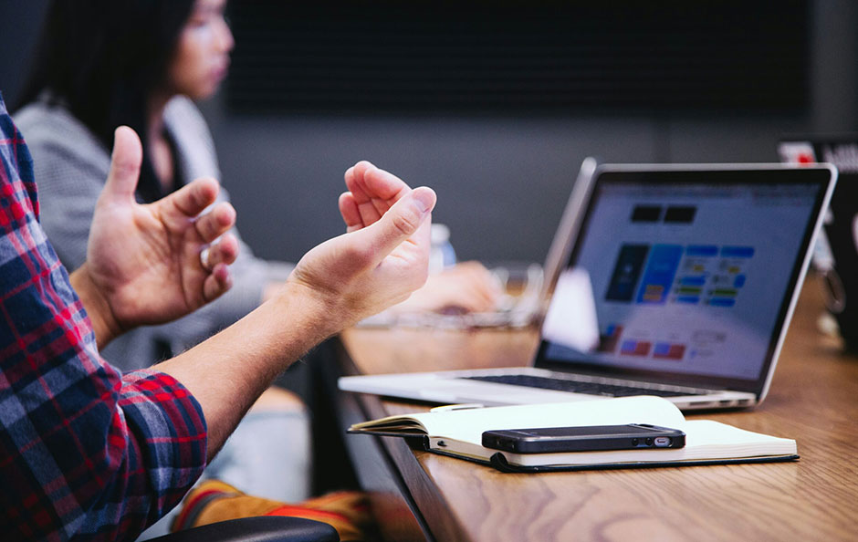 A person in a plaid shirt gesturing with their hands while talking in front of a laptop.
