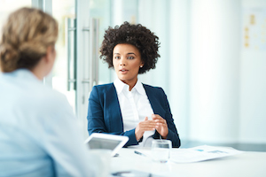 A professional woman with an afro hairstyle speaking to another person across a desk in a bright office.