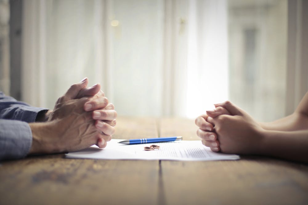 Two people sitting opposite each other at a table with clasped hands, appearing to be in mediation or disagreement.