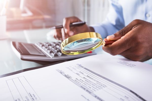A person using a magnifying glass to inspect financial documents with a calculator nearby.