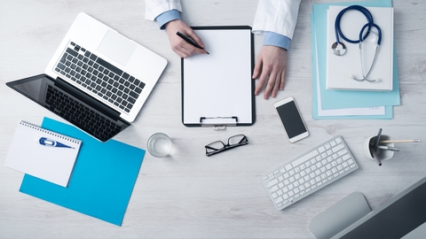 An overhead view of a doctor's desk with a laptop, clipboard, phone, and medical equipment.