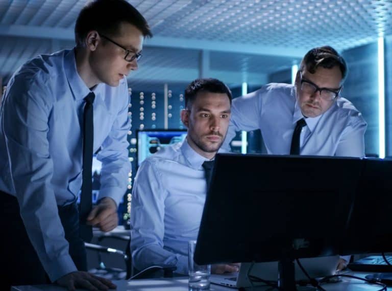 Three businessmen in a modern office looking intently at a computer screen.