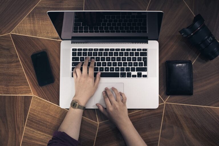 An overhead view of a person's hands typing on a laptop on a wooden floor.