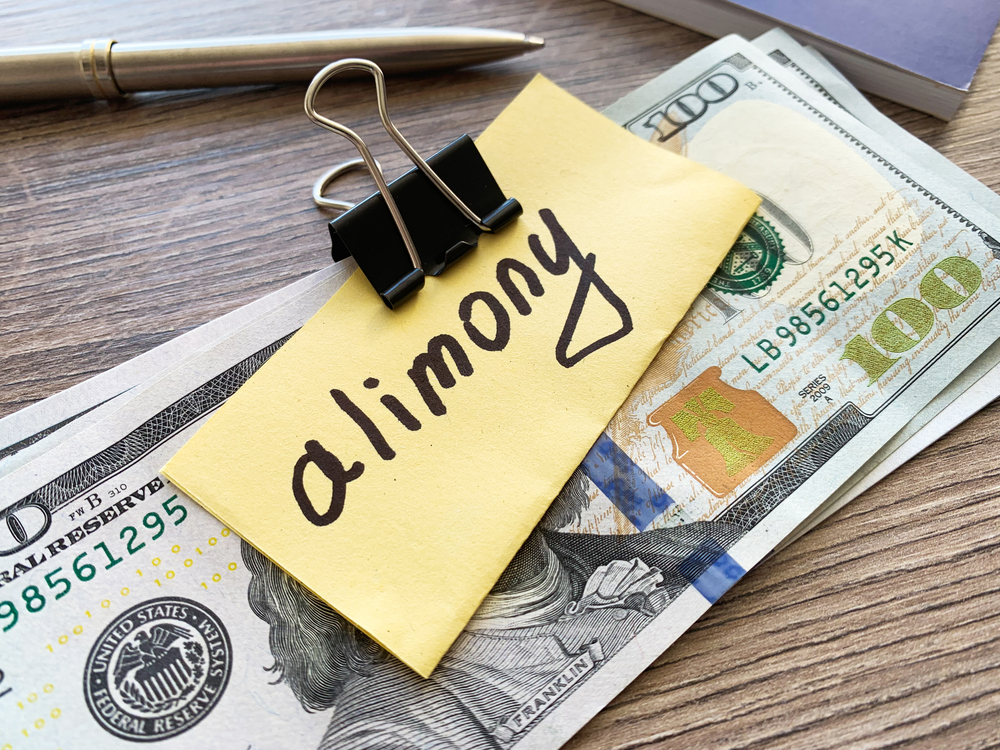 A yellow sticky note with the word 'alimony' handwritten on it, clipped to US dollar bills.
