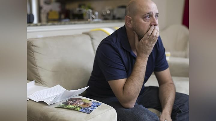 A worried man sits on a couch, covering his mouth with his hand, with photos on the cushion beside him.