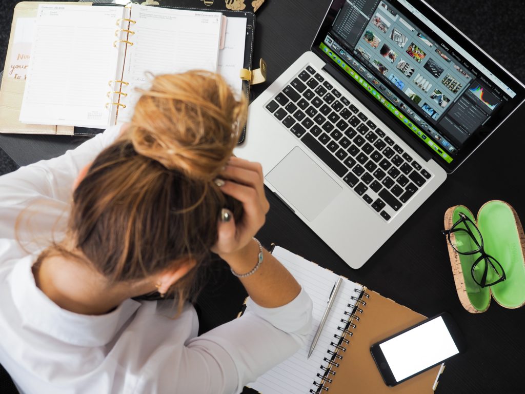 A frustrated person with their hair in a bun, holding their head while sitting at a desk with a laptop.
