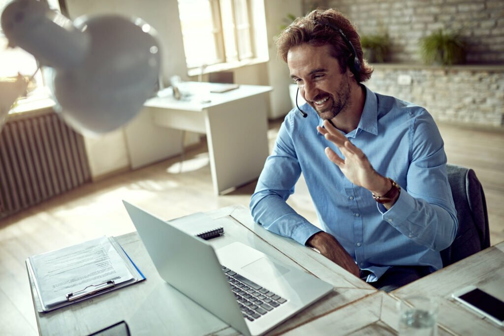 A happy man with a headset waving during a video call on his laptop.