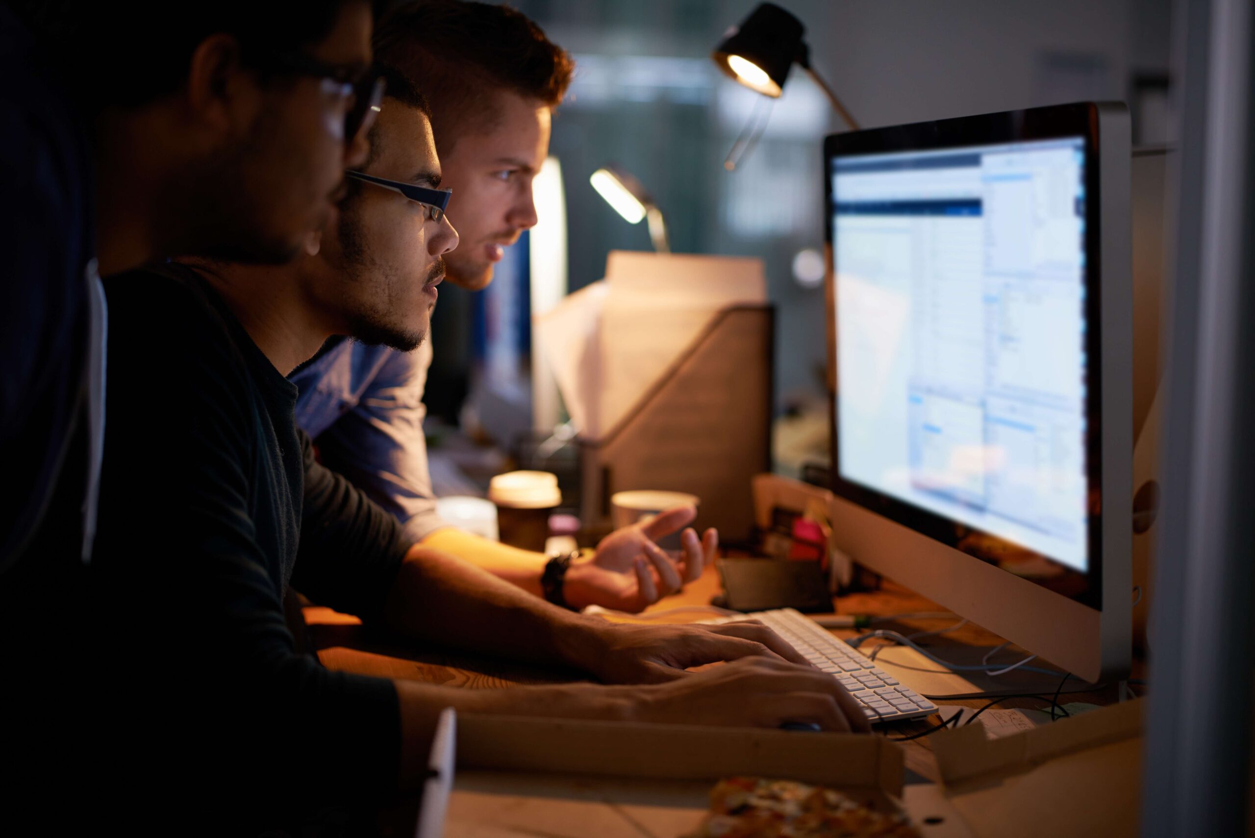 A team of people gathered around a computer screen in a dark room, working together.