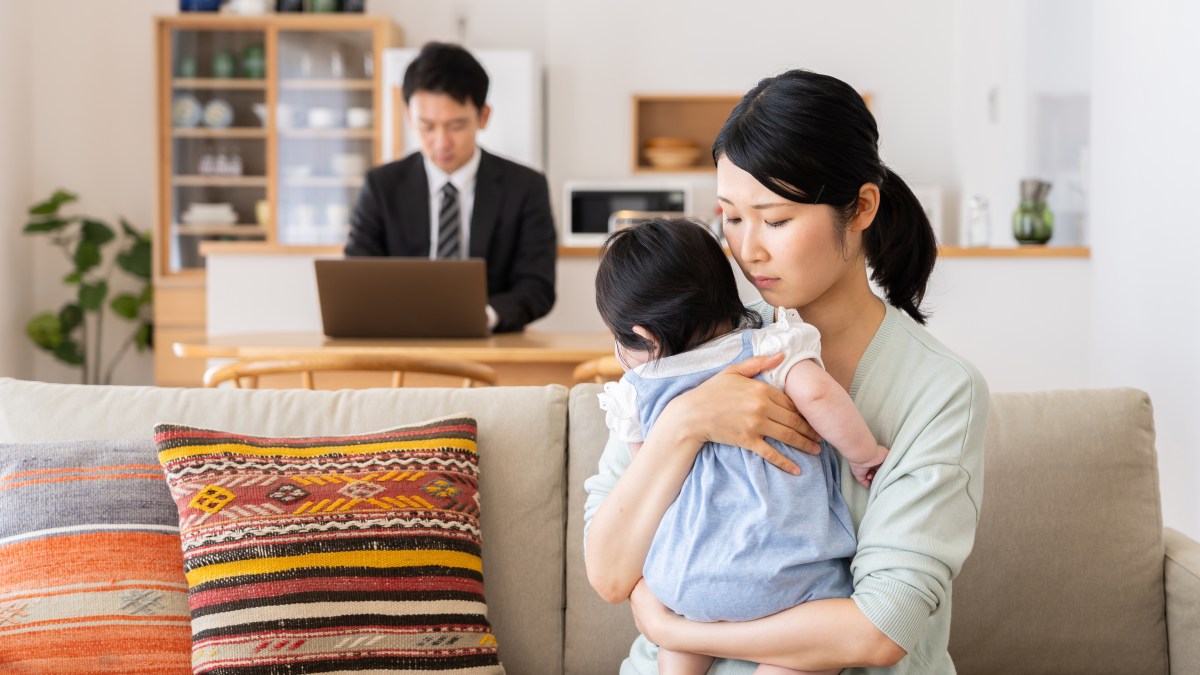 A sad mother holding her child on the couch while her husband works on a laptop in the background.