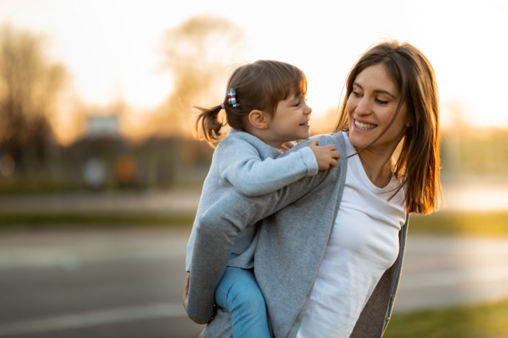 A smiling mother giving her young daughter a piggyback ride outdoors at sunset.