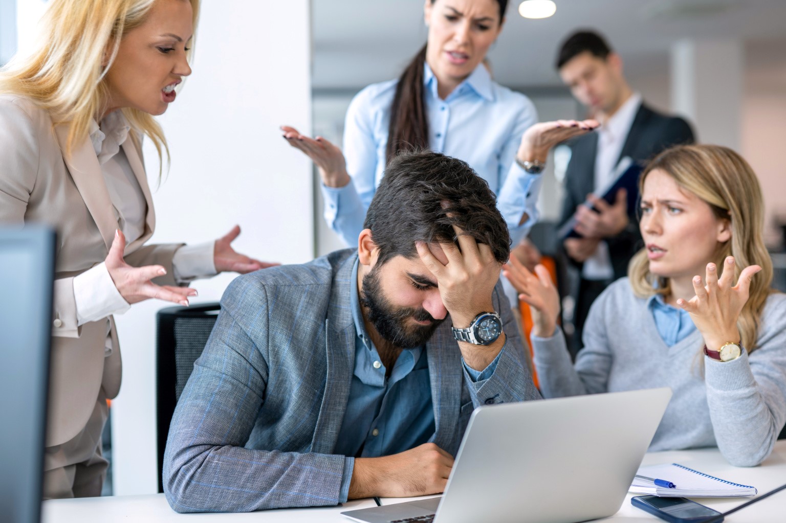 A stressed businessman holding his head at a table while colleagues argue around him.
