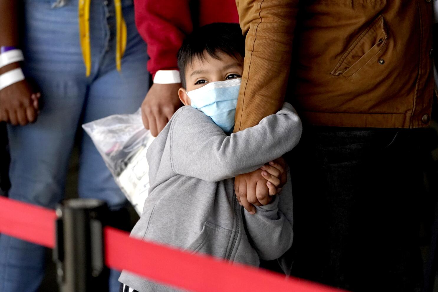 A young child wearing a face mask fearfully hugs an adult's leg.