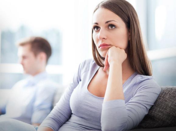 A woman sits on a couch looking worried, with a man blurred in the background.
