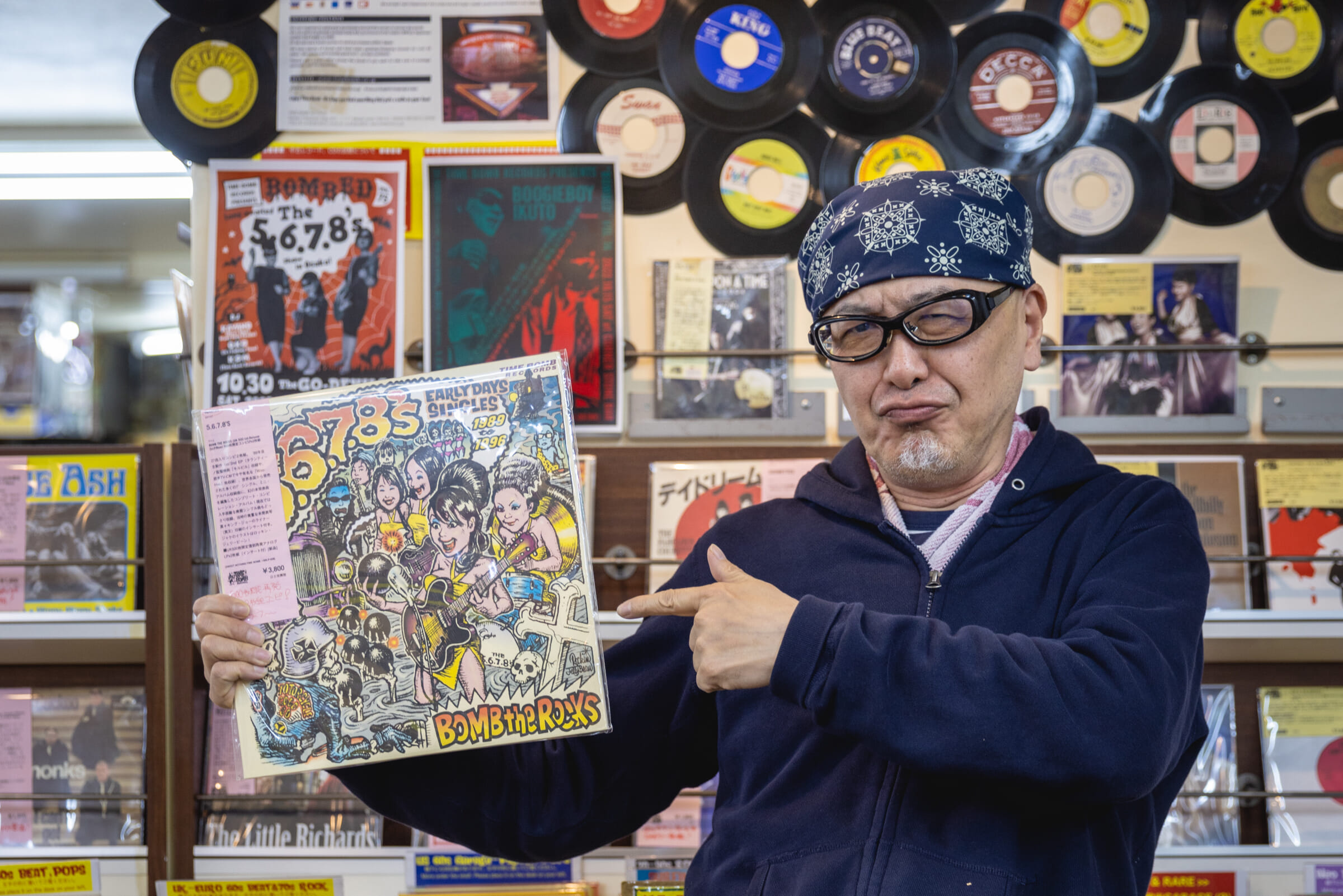 A record store owner with a bandana and glasses pointing to a rare record he is holding.