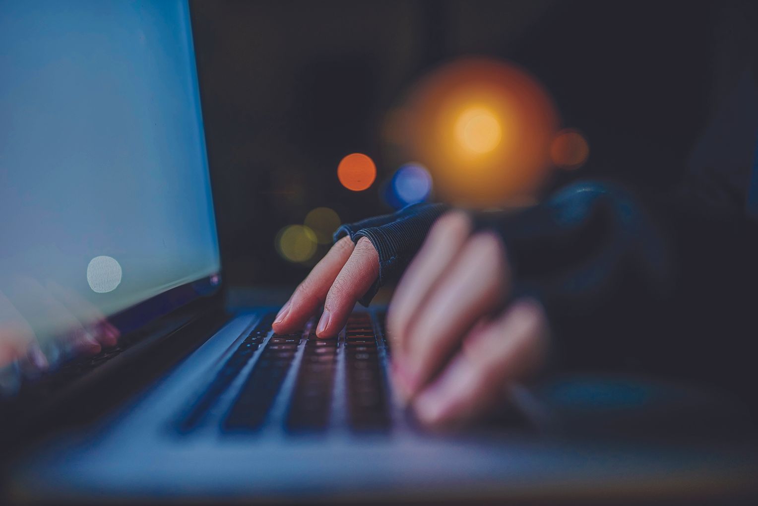 Close-up of hands typing on a laptop keyboard in a dark room with bokeh lights in the background.