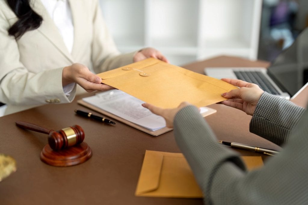 A person in a suit handing a brown envelope to another person over a desk with a gavel.