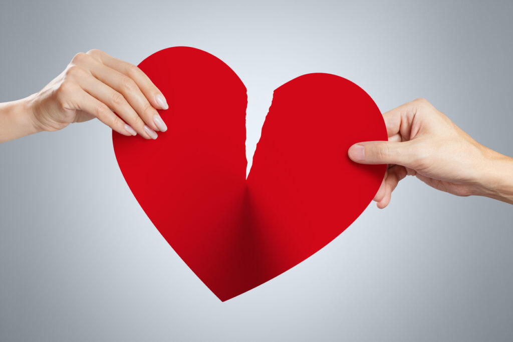 Two hands holding a torn red paper heart against a gray background.