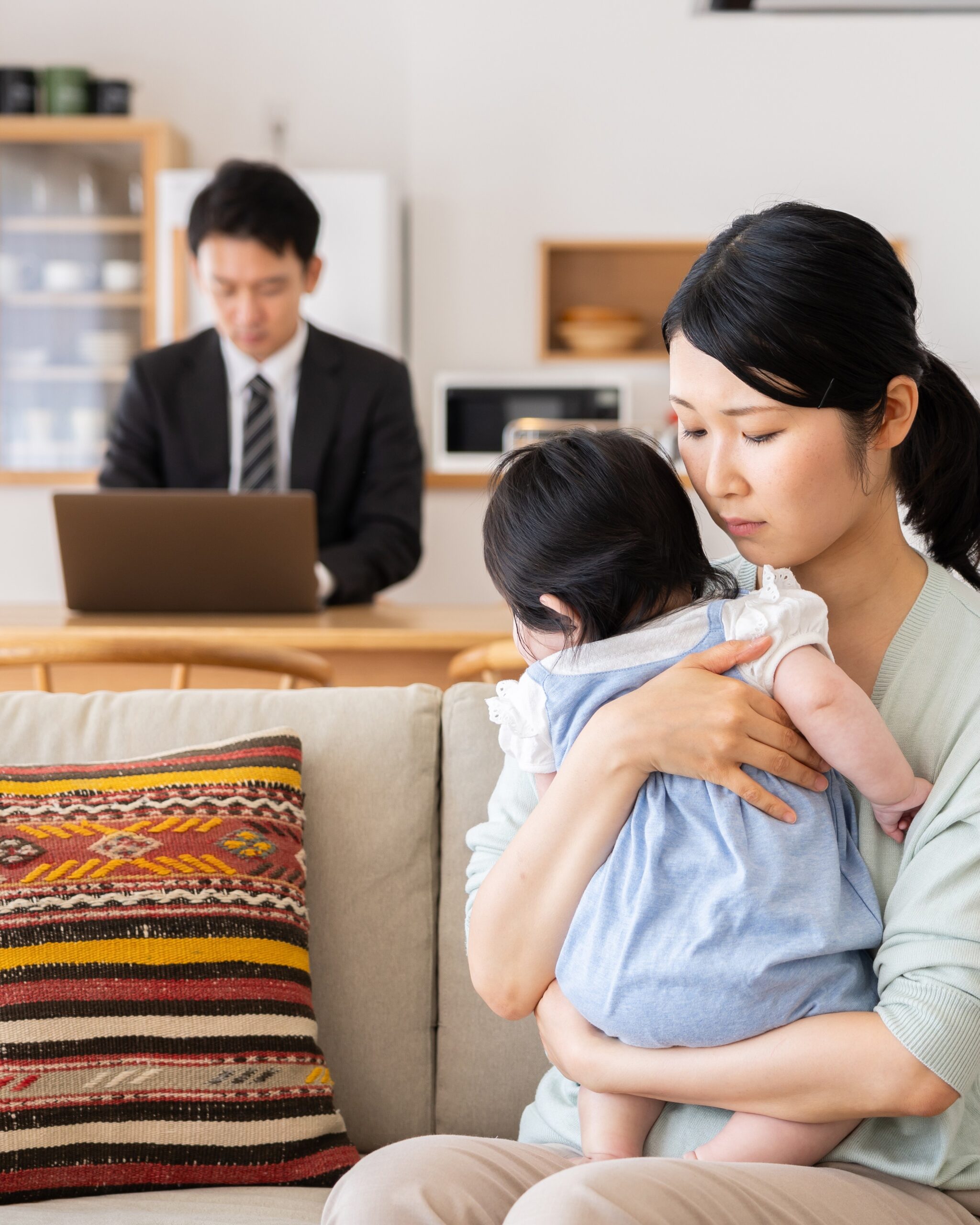 A sad mother holding her child on the couch while her husband works on a laptop in the background.